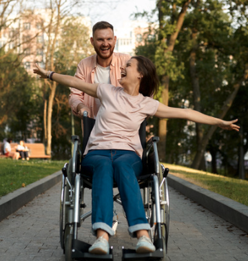 Woman in a wheelchair being pushed through a park by a man. Both look very happy and the woman has her arms outstretched to the side.d 