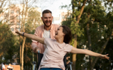 Woman in a wheelchair being pushed through a park by a man. Both look very happy and the woman has her arms outstretched to the side.d 
