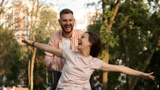 Woman in a wheelchair being pushed through a park by a man. Both look very happy and the woman has her arms outstretched to the side.d 