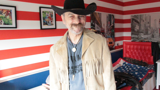 Trevor stood in his bedroom with a cowboy hat on. The wall paper behind him is red and white horizontal stripes.