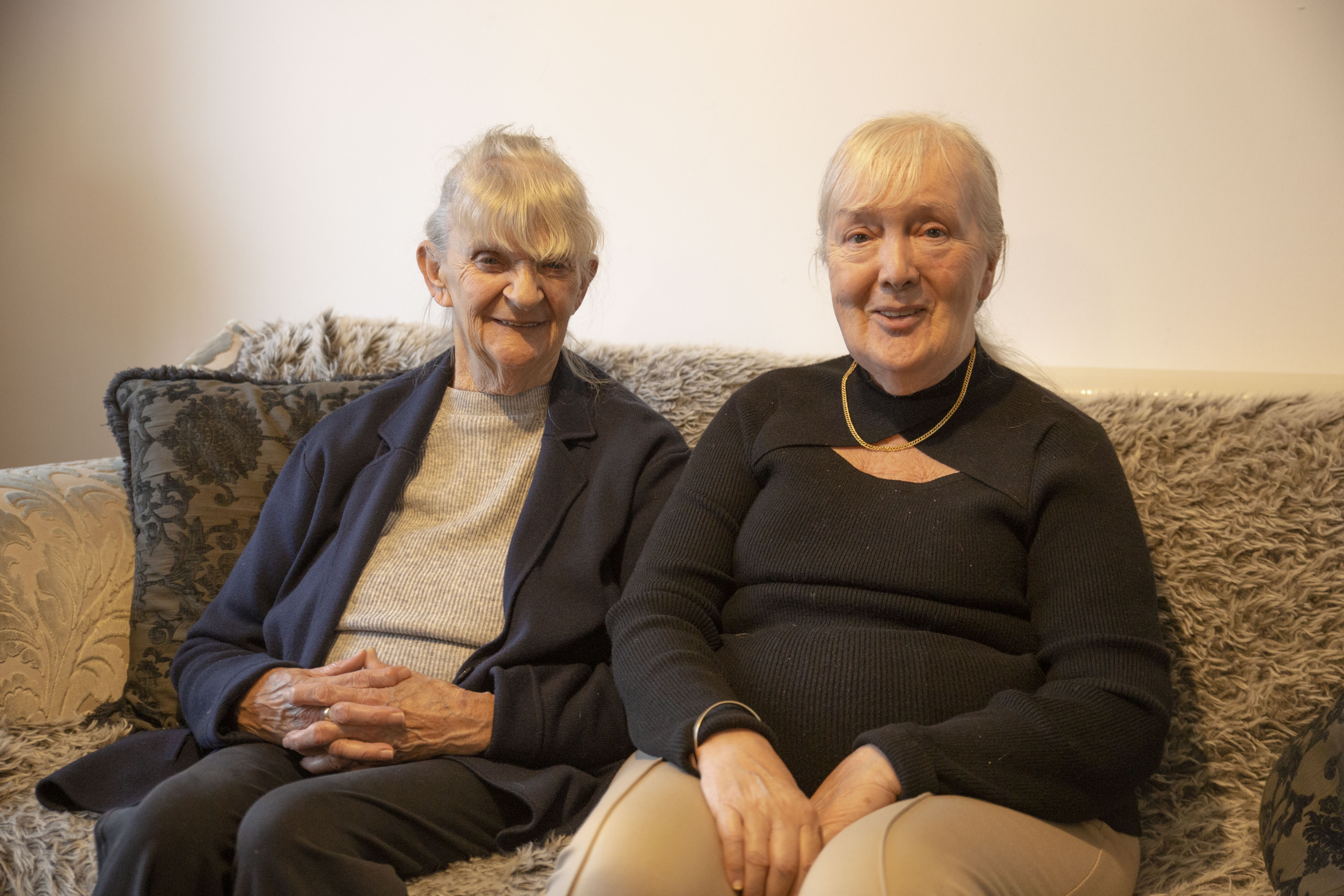 Two older ladies sat on a sofa smiling. 