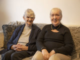 Two older ladies sat on a sofa smiling. 