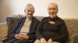 Two older ladies sat on a sofa smiling. 