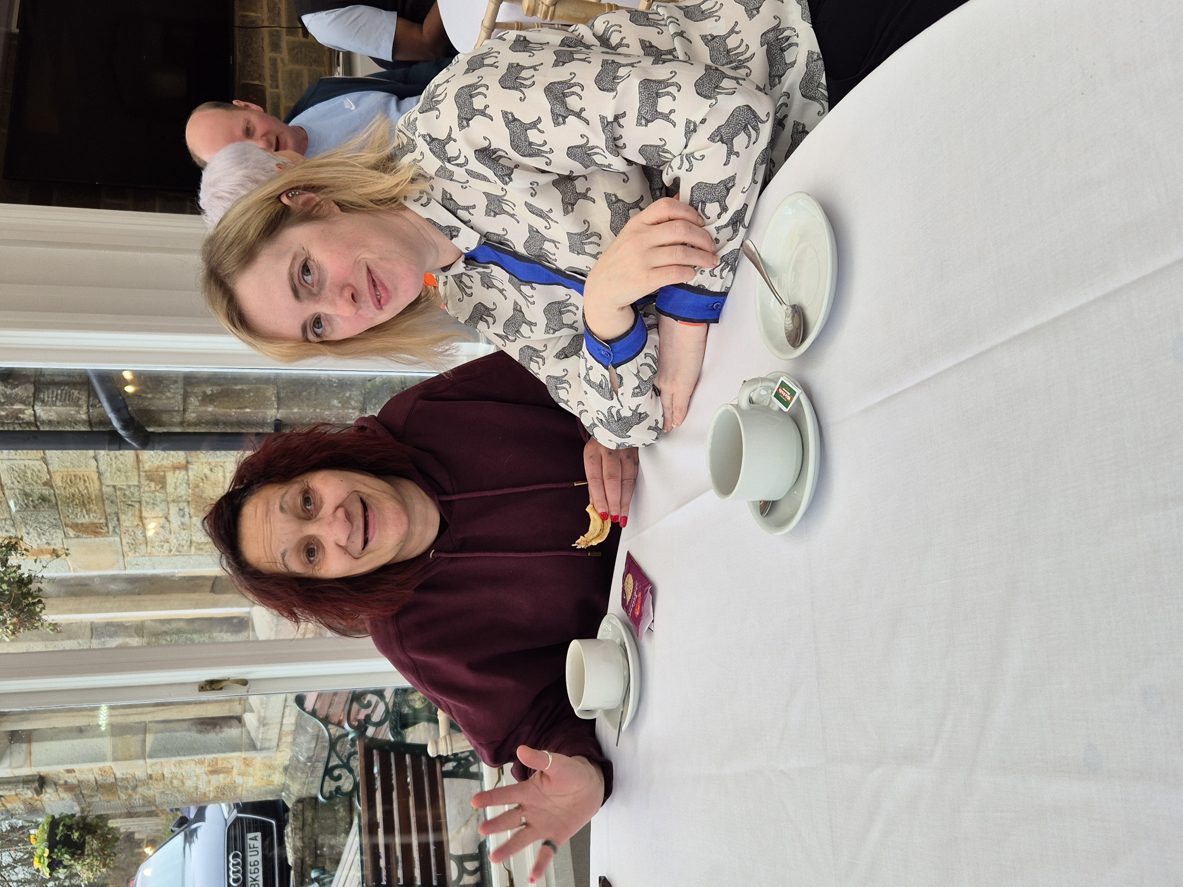 Two women sat down having tea while looking at the camera and smiling