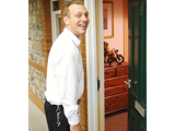  A man at an opened front door holding a suitcase and smiling.  
