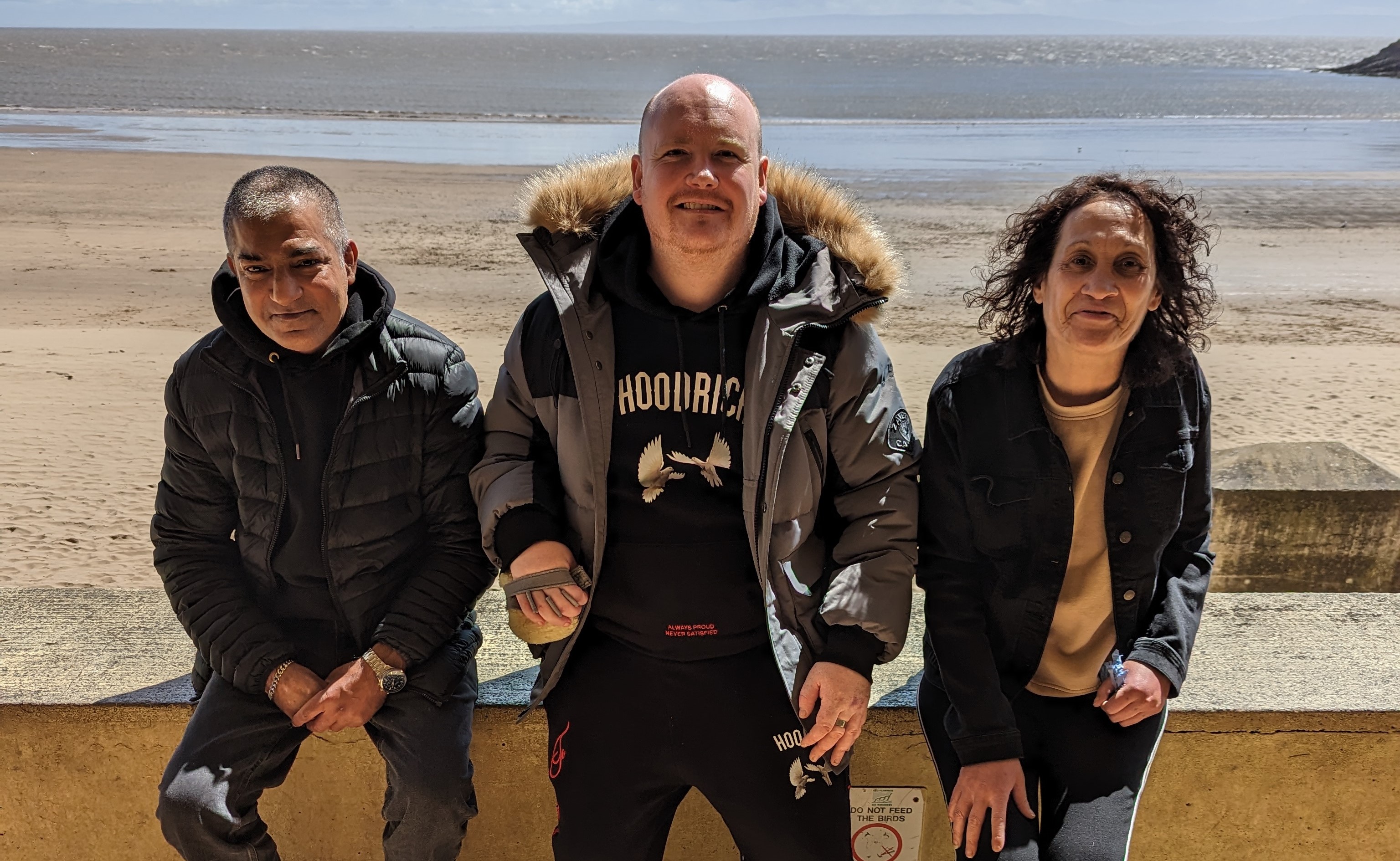 Three smiling residents of Ty Cornel, two men and a woman, sitting on a wall with a view of the beach and the ocean behind them. 