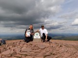 A man and a woman stood next to the Pen Y Fan sign on top of the mountain. 